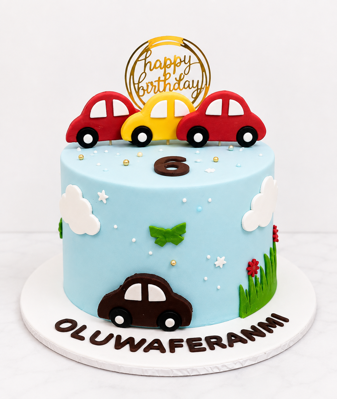 Car Themed Kids Birthday Cake Sky blue fondant kids birthday cake with red and yellow toy car toppers, clouds, and grass detailing by Cakes by Teez in Abuja