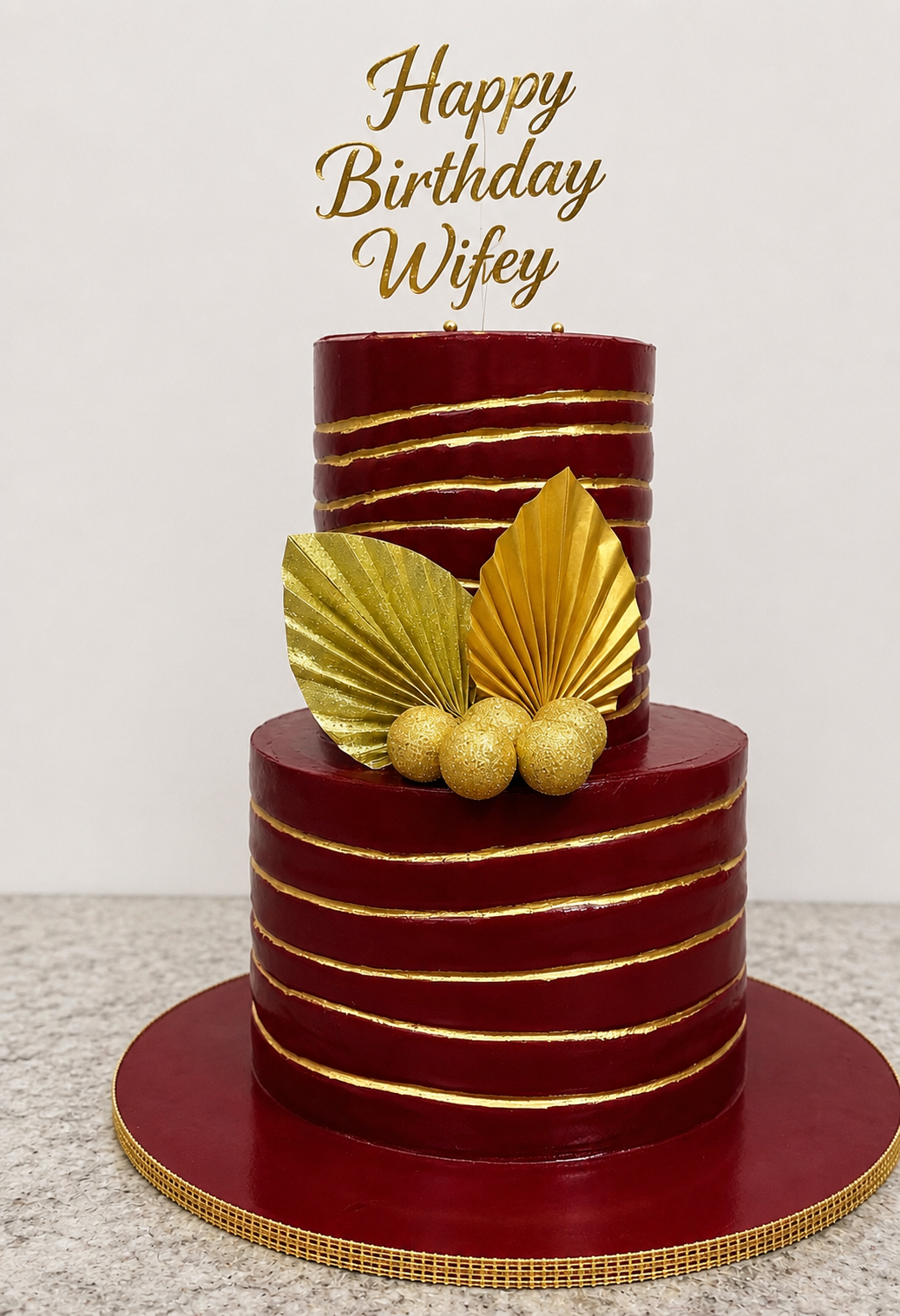 Two-tier burgundy buttercream birthday cake with gold leaf line detailing and gold fondant leaf and sphere accents by Cakes by Teez in Abuja