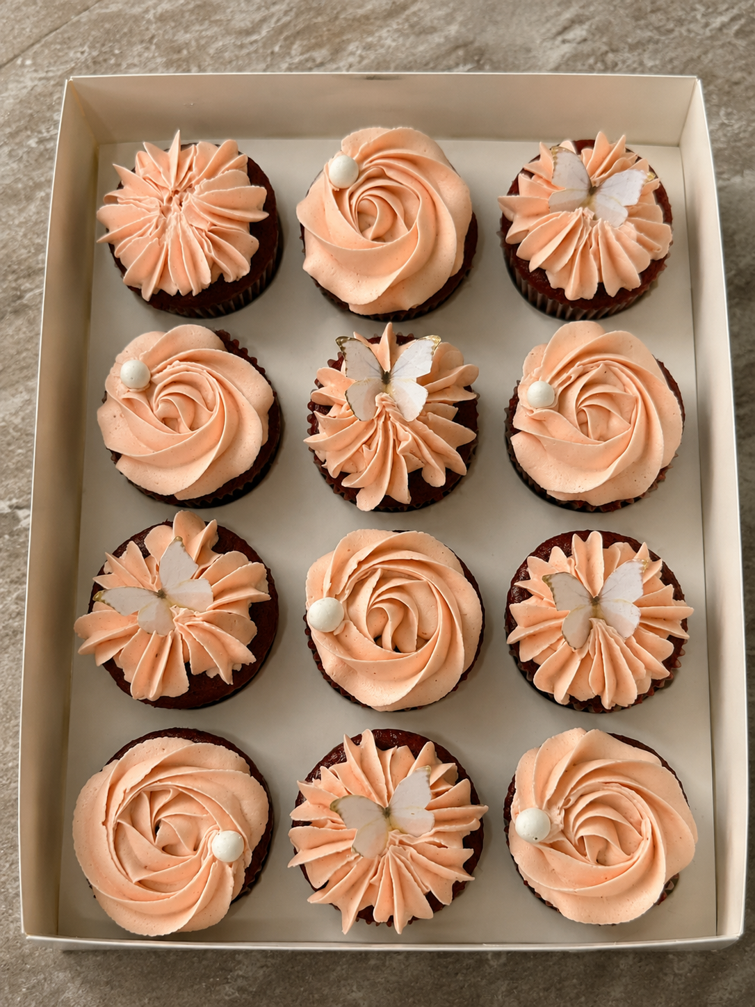 Box of twelve cupcakes with peach buttercream rose swirls and edible butterfly toppers by Cakes by Teez in Abuja