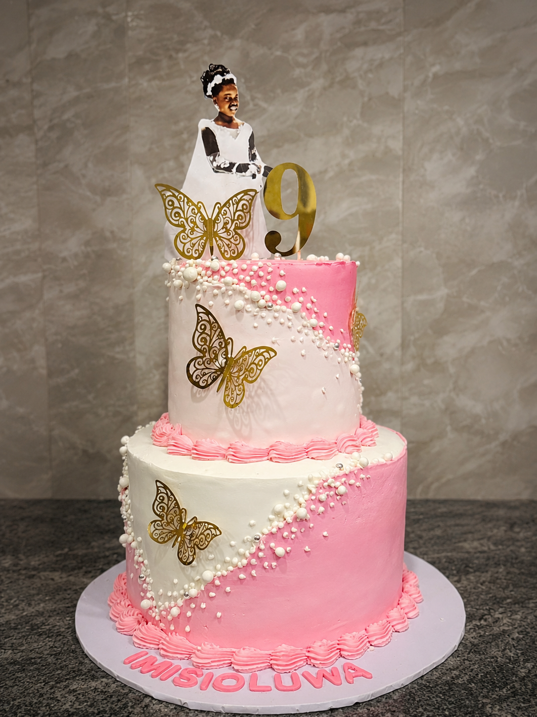 Two-tier pink and white buttercream birthday cake with gold butterfly accents, pearl details, and a personalised photo topper by Cakes by Teez in Abuja