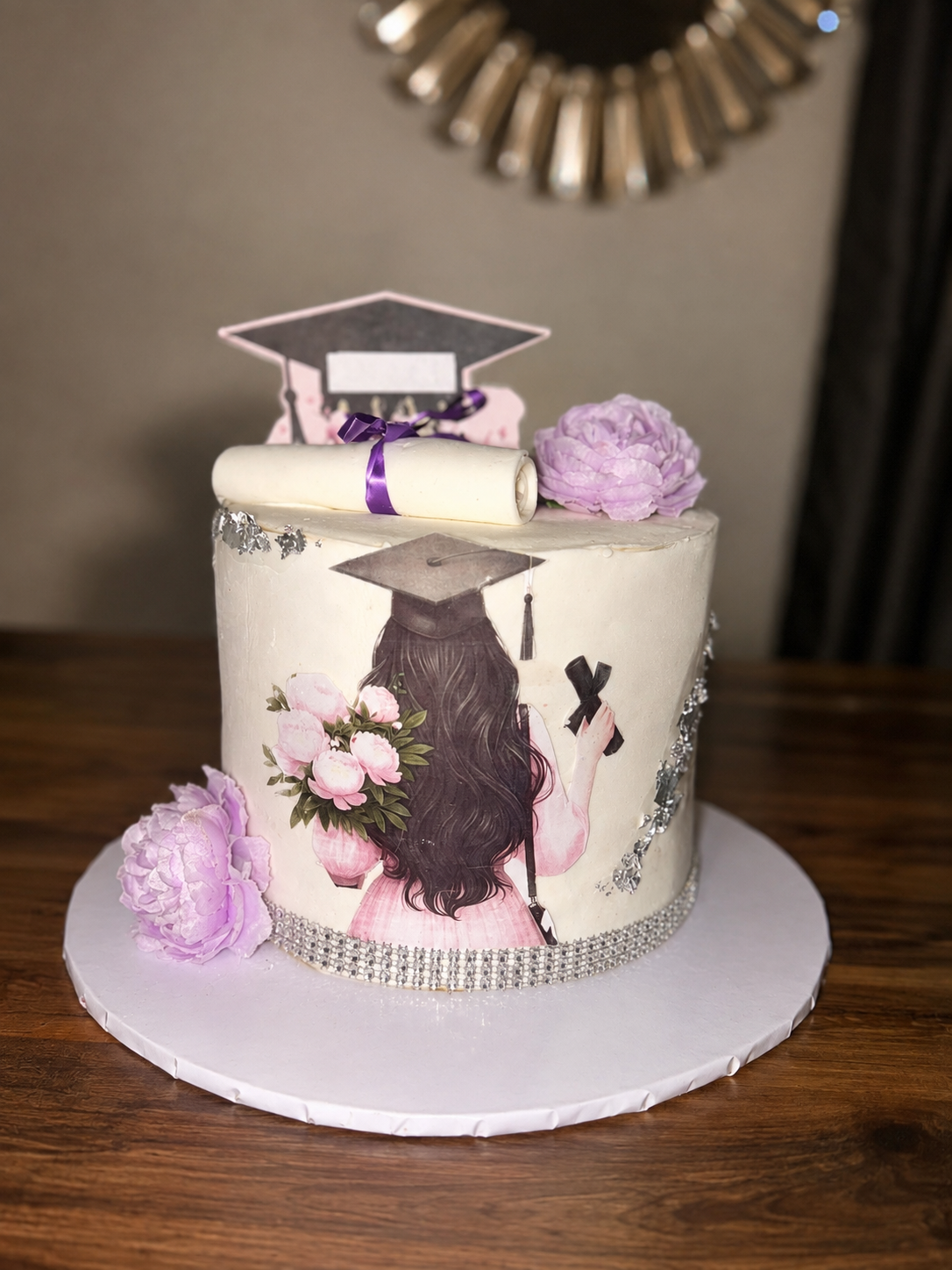 Ivory buttercream graduation cake with an edible illustrated print of a graduate, fresh lilac peonies, and silver leaf accents by Cakes by Teez in Abuja