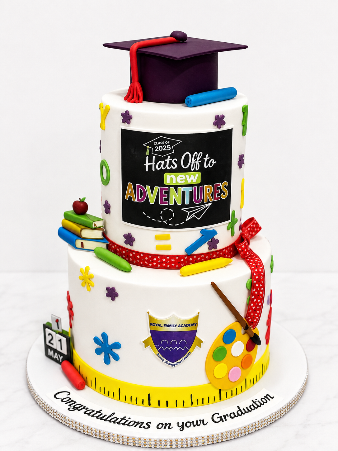 Two-tier white fondant school graduation cake with a graduation cap, books, paint palette, and a school crest topper by Cakes by Teez in Abuja